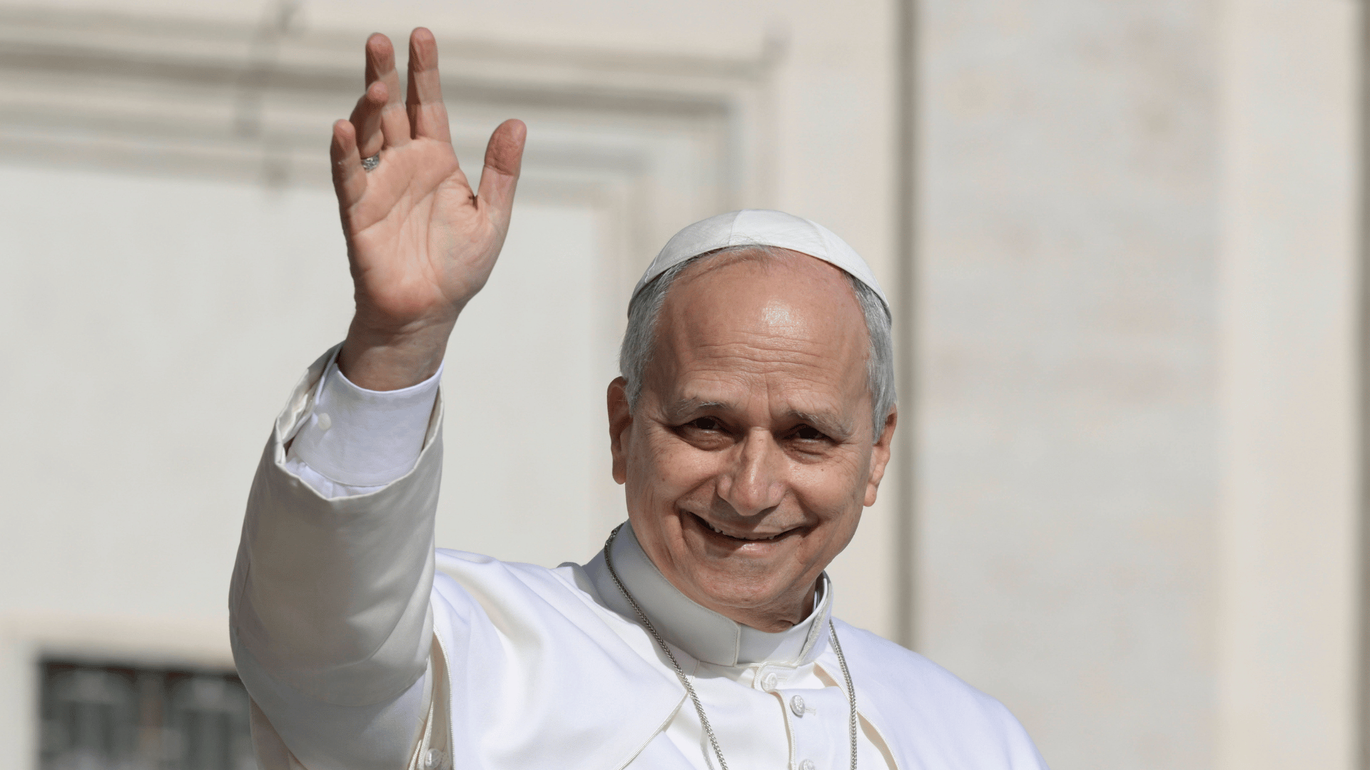 A imagem ilustra o Papa Leão XIV com suas habituais vestes pontifícias, sorrindo e com a mão direita erguida em sinal de aceno.