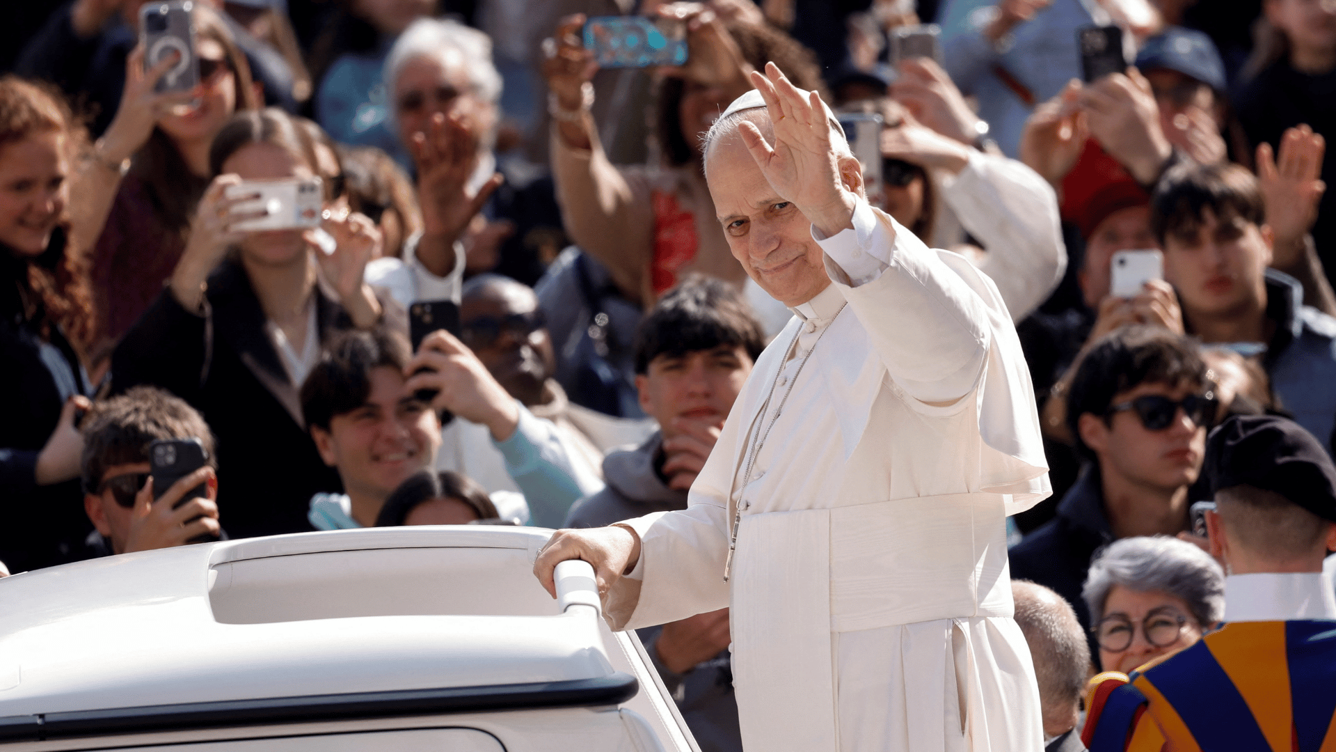 A imagem ilustra o Papa Leão XIV com suas habituais vestes pontifícias, no papamóvel, com o braço esquerdo erguido em gesto de aceno e uma multidão de fiéis ao fundo.