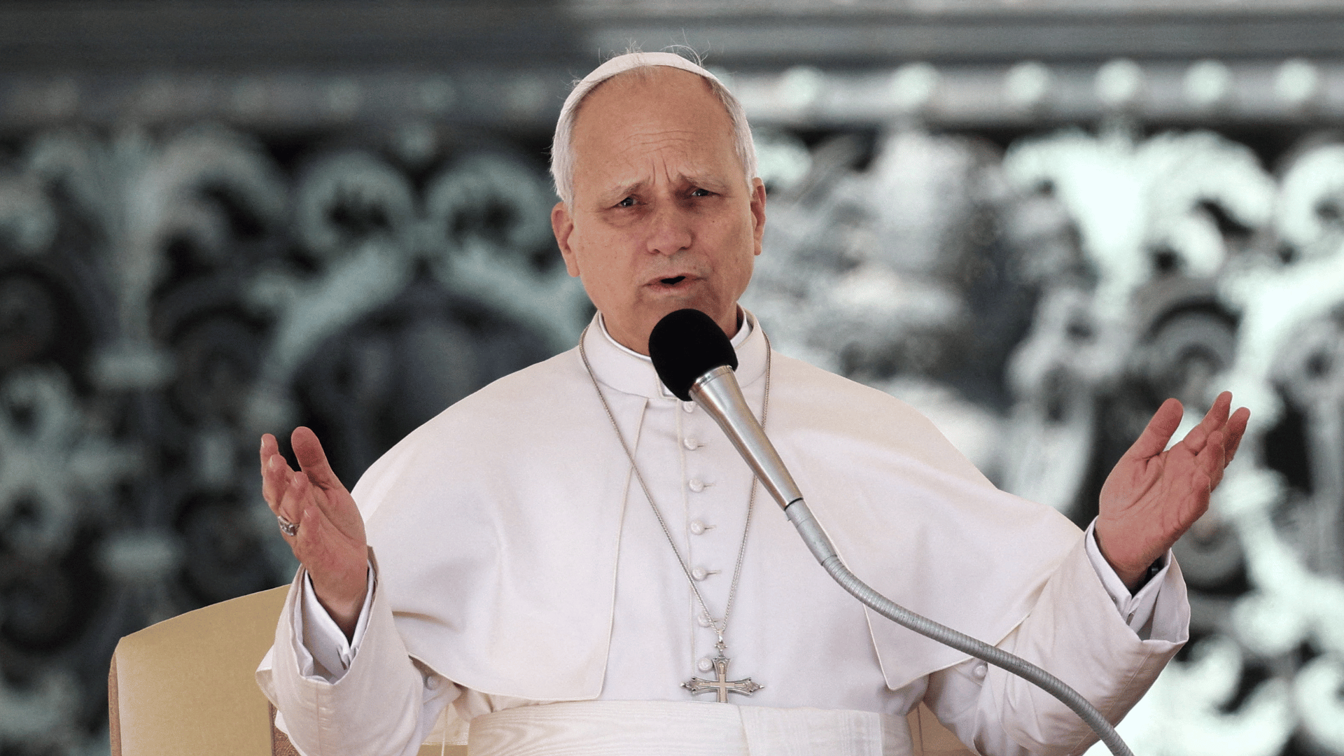 Papa vestido de branco fala ao microfone durante catequese, com as mãos abertas em gesto de explicação, em ambiente externo no Vaticano.
