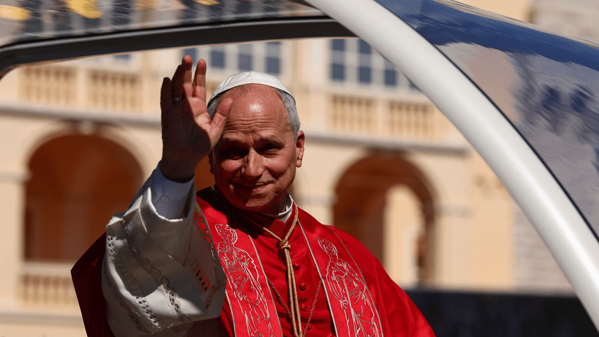 Papa Leão XIV Líder acena para fiéis durante deslocamento em veículo aberto, vestindo paramentos vermelhos e solidéu, em Mônaco.