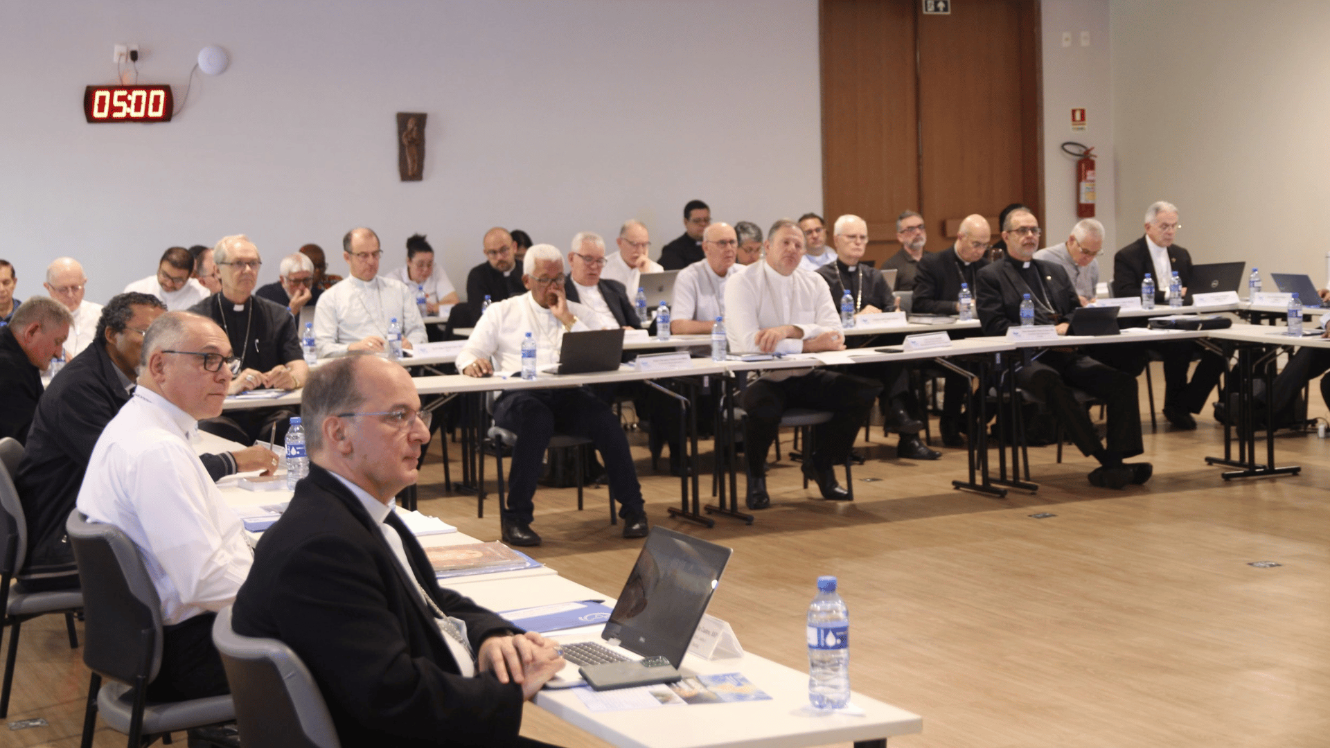 A imagem ilustra um grupo de bispos sentados em fileiras durante uma reunião