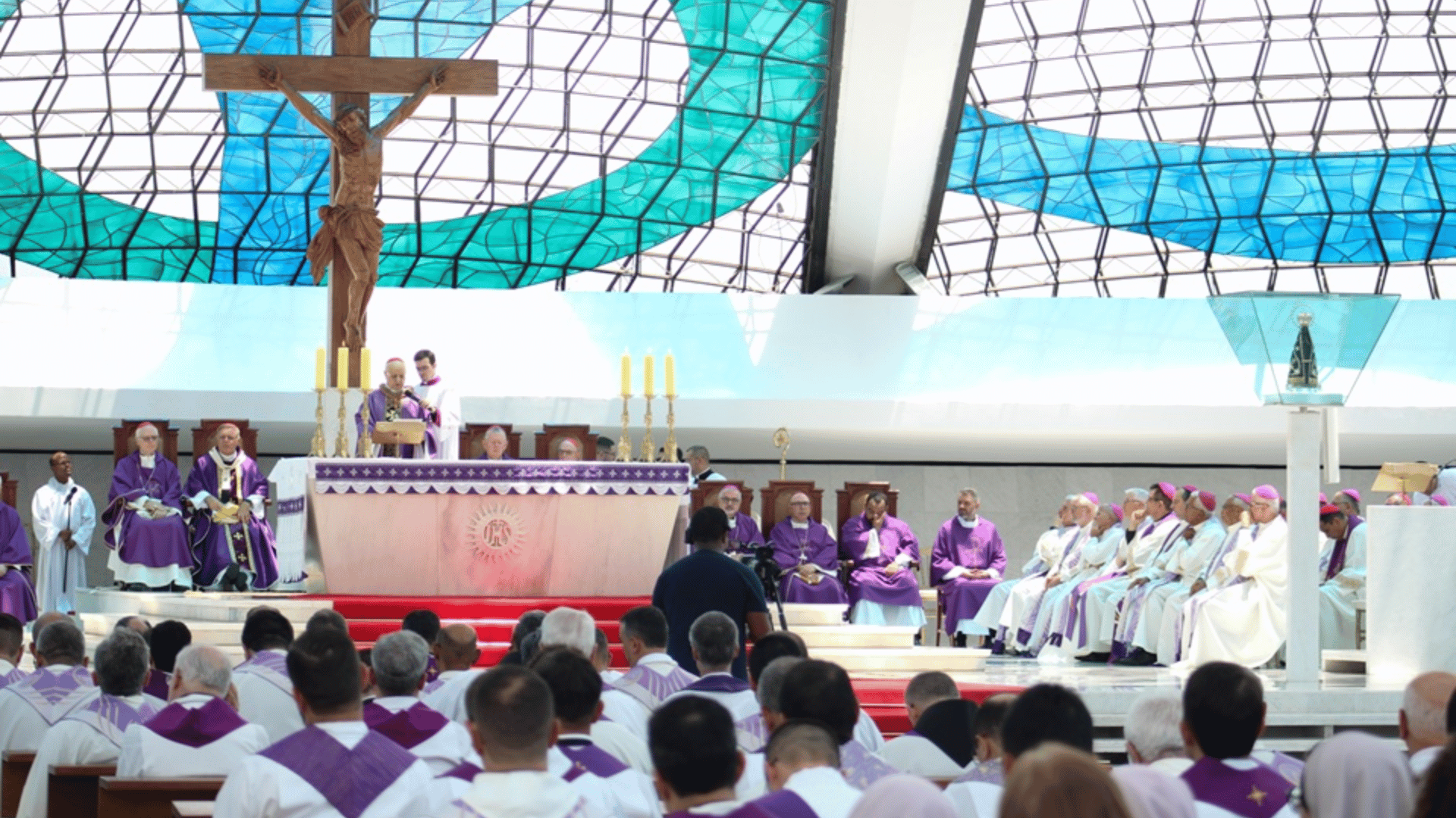 Celebração da Santa Missa em igreja, com bispos e sacerdotes vestindo paramentos roxos reunidos ao redor do altar, grande crucifixo suspenso ao centro e vitral azul ao fundo, com fiéis participando da celebração.