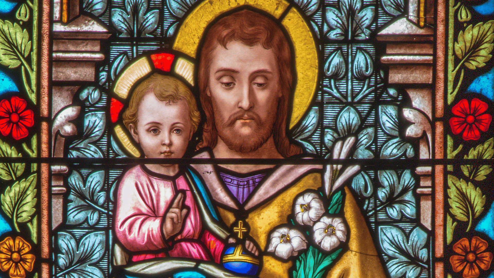 Vitral religioso representando São José com o Menino Jesus nos braços, ambos com auréolas, com lírios brancos ao lado e fundo decorado com flores coloridas