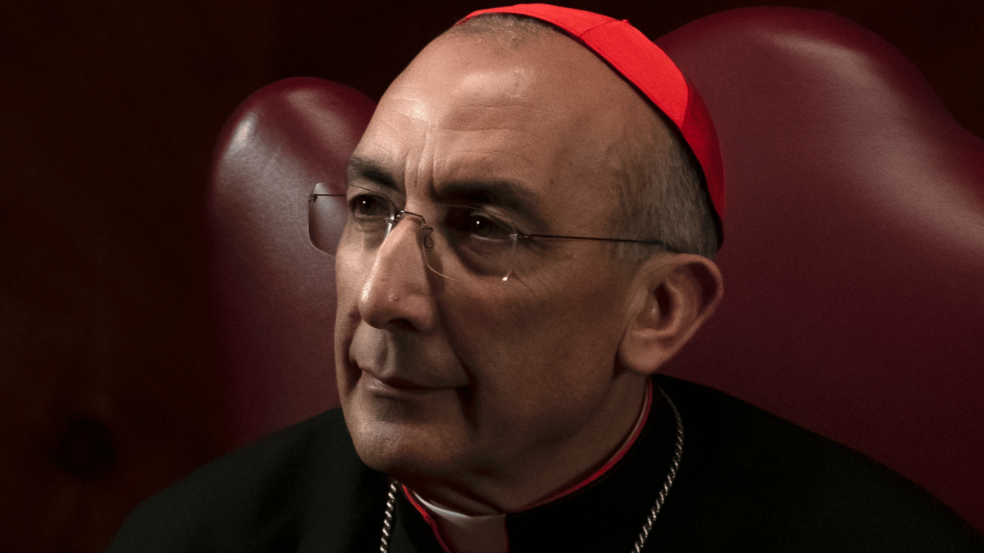 Cardeal Reina usando solidéu vermelho e óculos, aparece sentado em uma cadeira vermelha