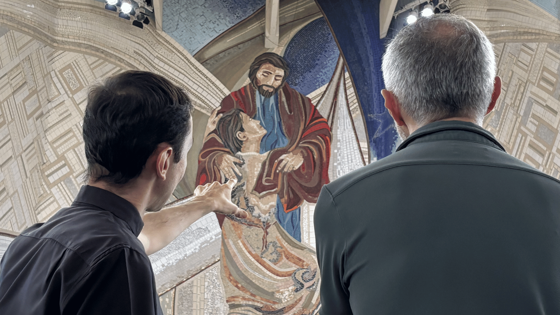 A imagem ilustra dois padres, de costas. O padre do lado esquerdo aponta, com a mão esquerda, para um mosaico que ambos contemplam