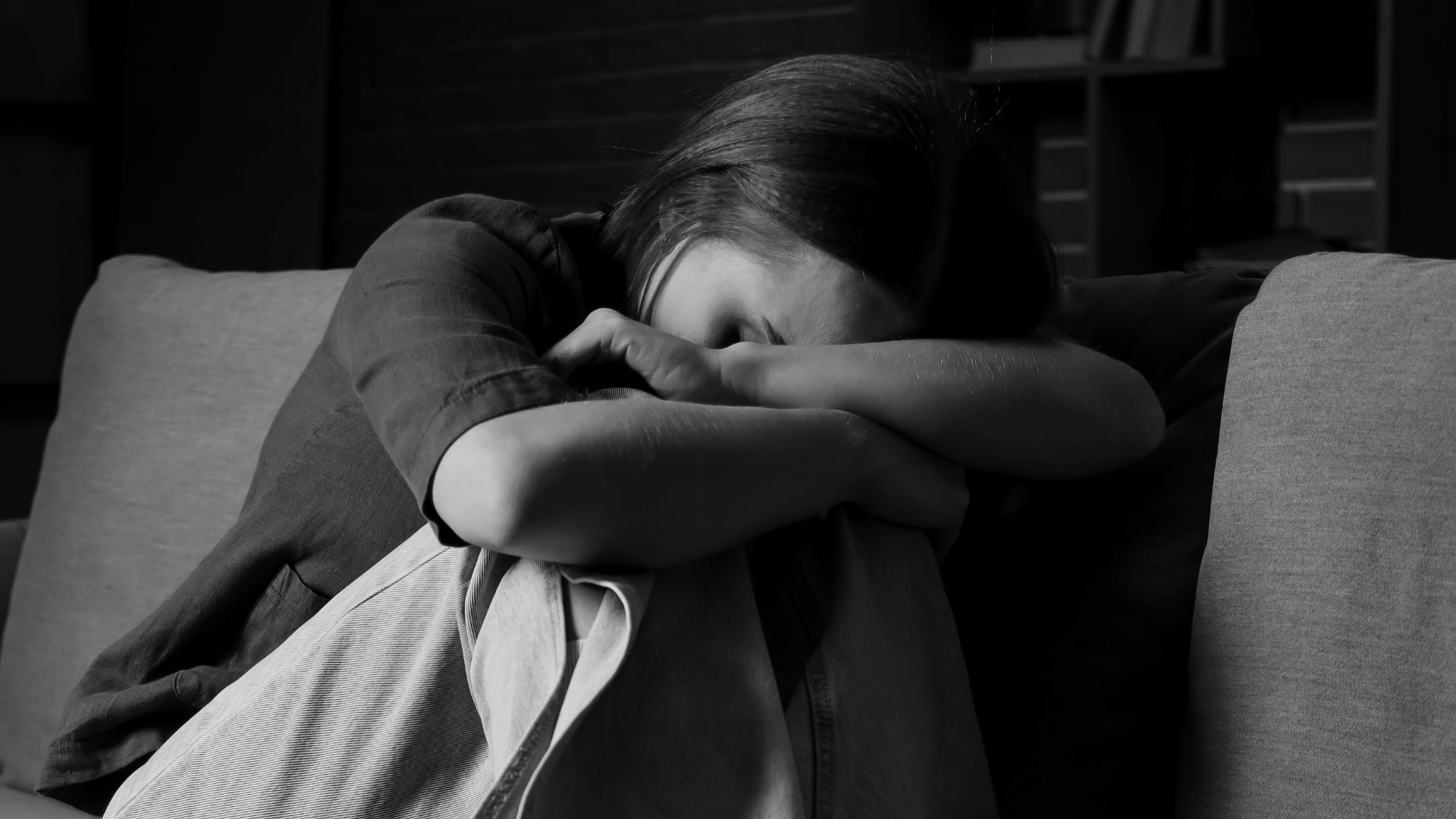 A imagem em preto e branco ilustra uma jovem sentada sobre um sofá, com as pernas recolhidas e os braços apoiados sobre os joelhos, escondendo seu rosto entre eles.