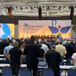 Participantes reunidos em auditório durante a 62ª Assembleia Geral dos Bispos do Brasil, voltados para o palco principal com painel temático e estrutura de iluminação.