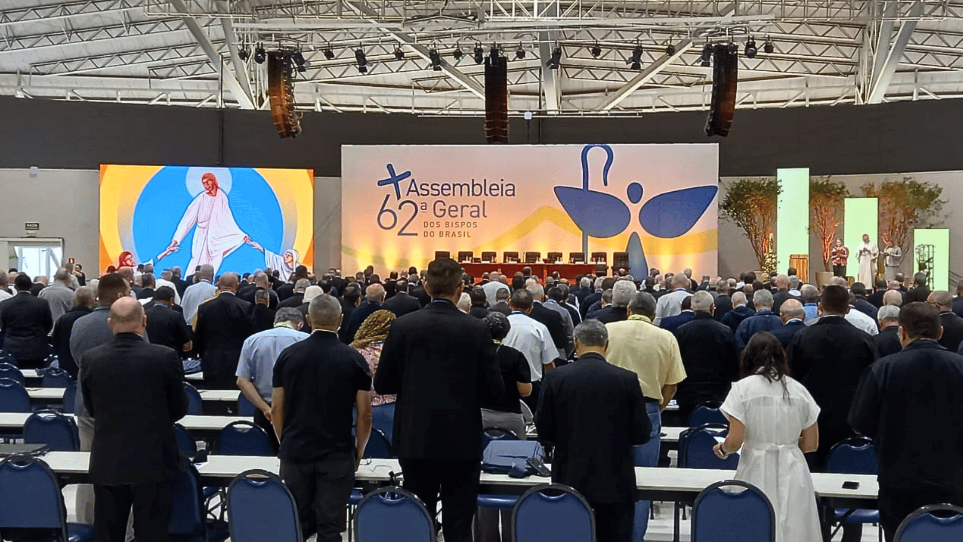 Participantes reunidos em auditório durante a 62ª Assembleia Geral dos Bispos do Brasil, voltados para o palco principal com painel temático e estrutura de iluminação.