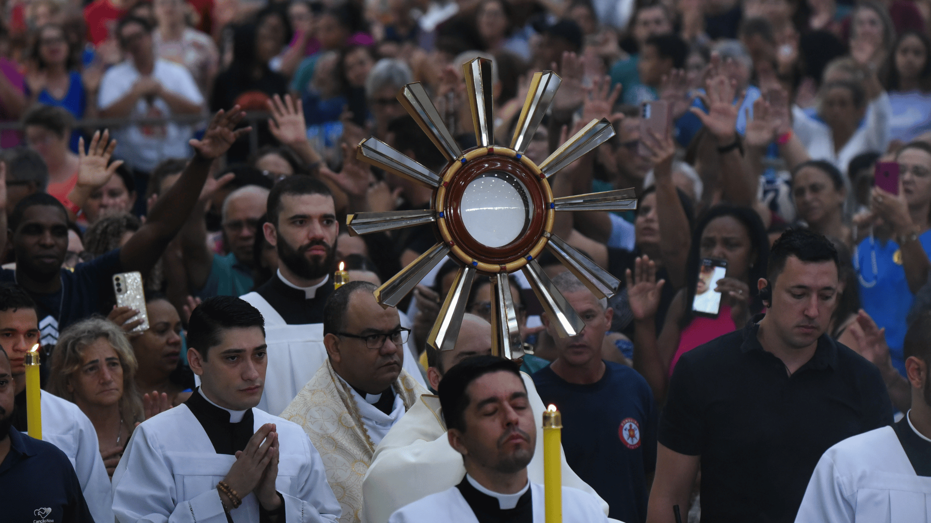 A imagem ilustra um padre carregando um ostensório com o Santíssimo Sacramento, em procissão, cercado por fiéis