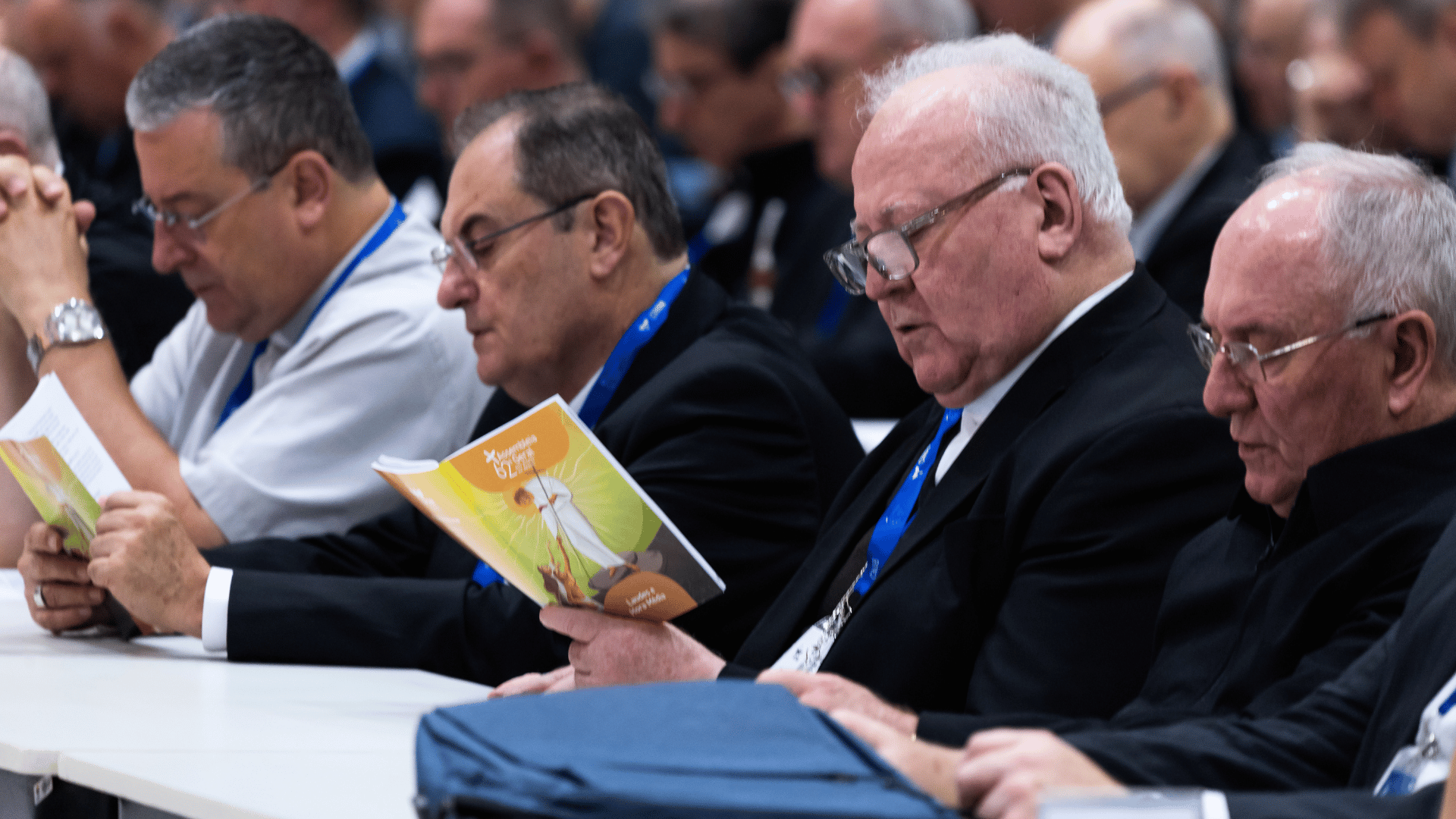 Bispos reunidos em assembleia, sentados em mesa, acompanham a programação enquanto leem material impresso durante encontro da Igreja no Brasil.