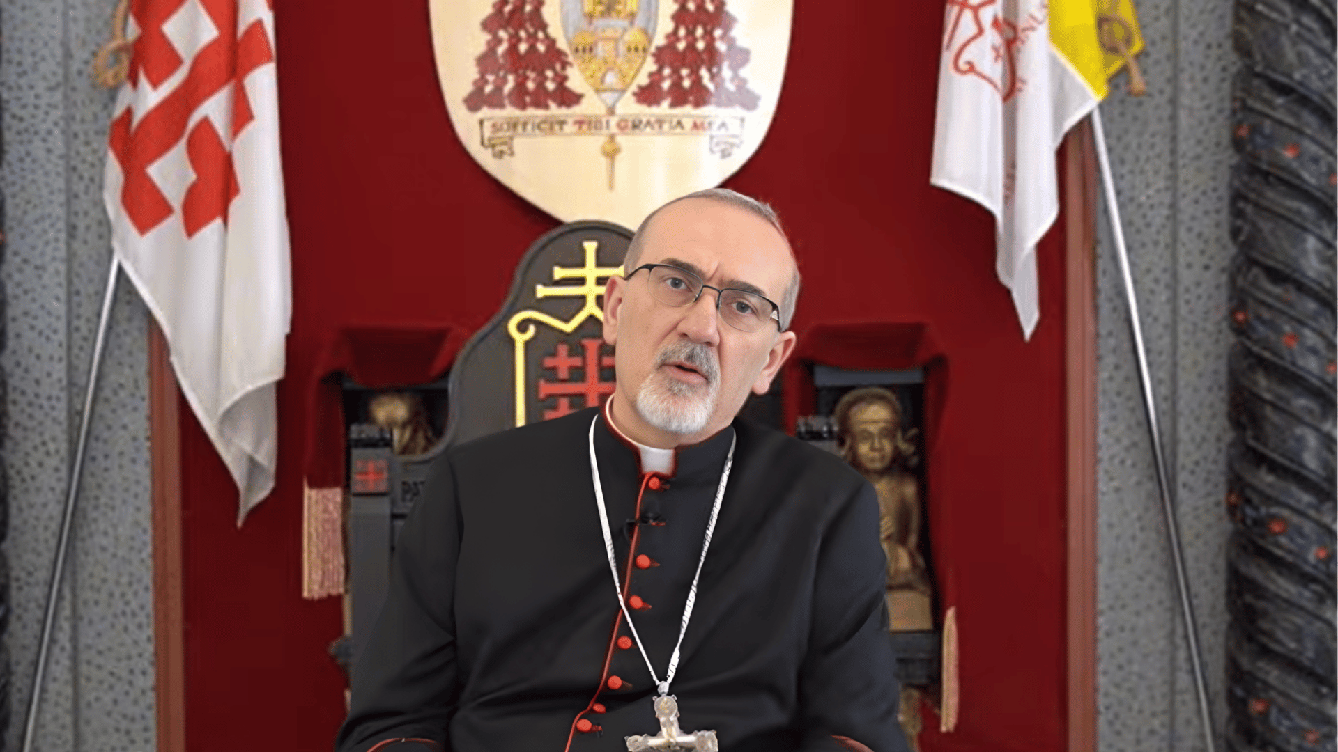 Cardeal Pierbattista Pizzaballa grava mensagem em vídeo sentado em cadeira oficial, diante de brasão e bandeiras institucionais.