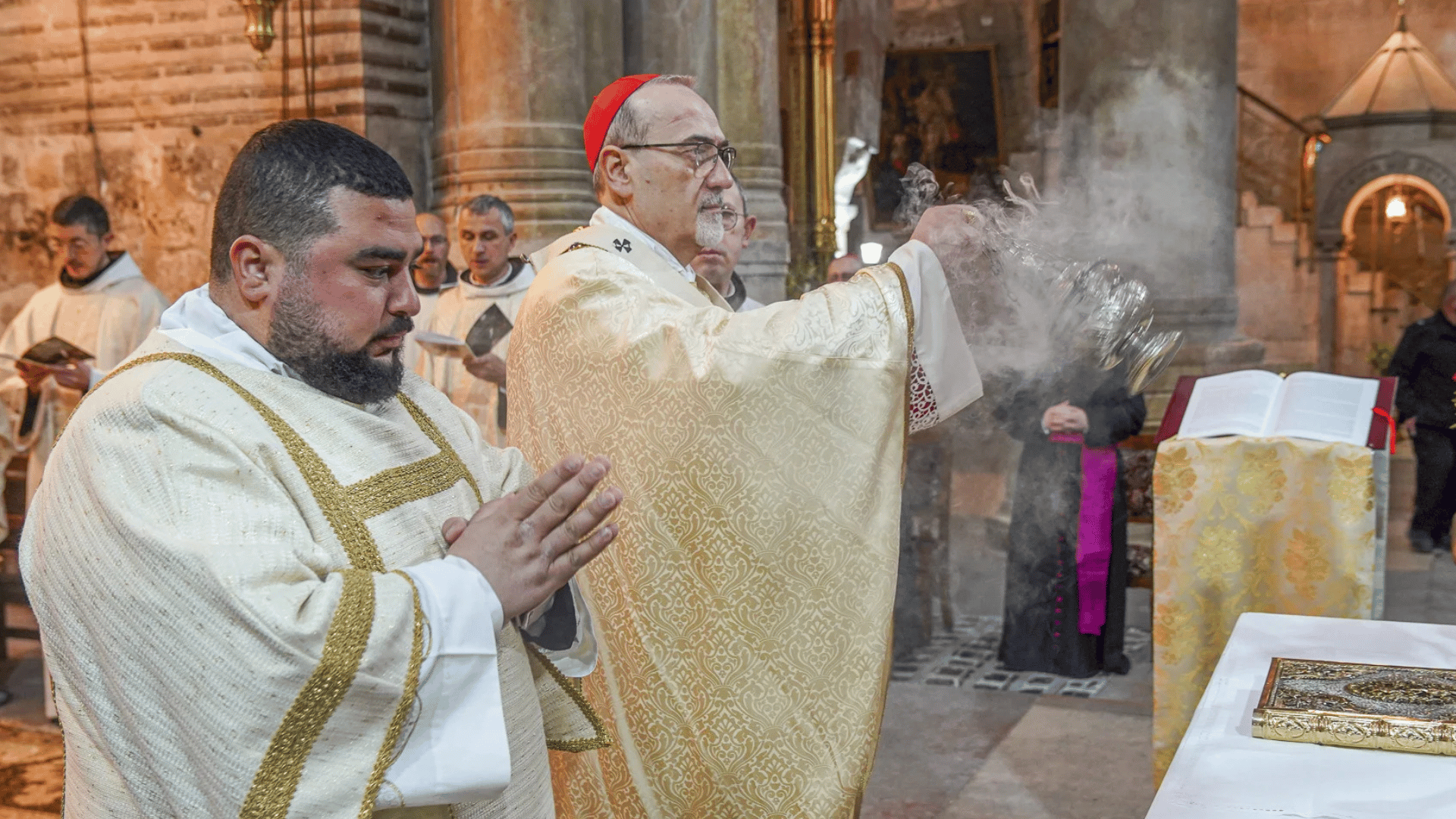 A imagem ilustra um cardeal da Igreja Católica, paramentado com vestes litúrgicas douradas, manuseando um turíbulo com um diácono à sua direita.