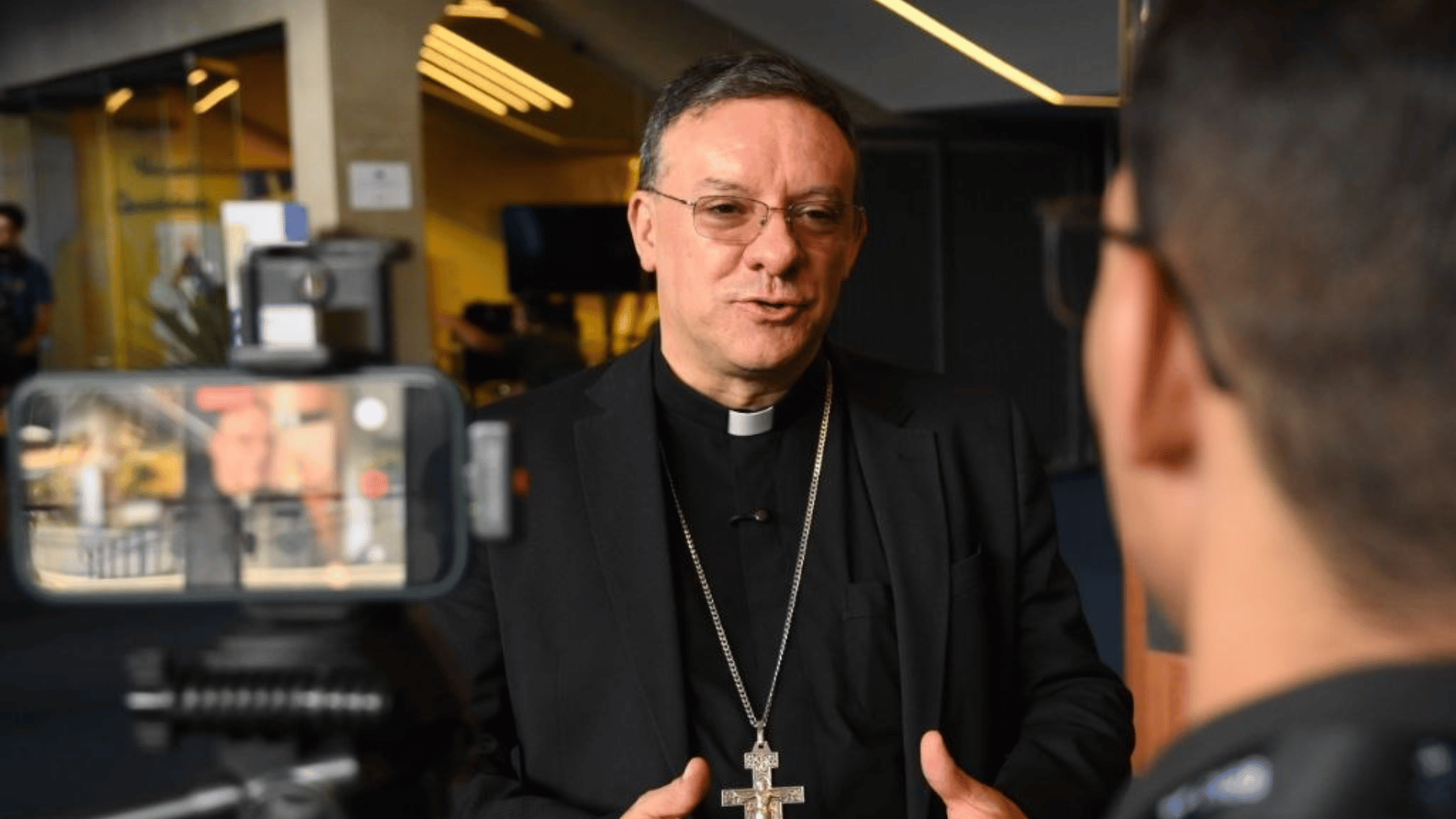 A imagem ilustra um bispo vestido com clérgima e blazer. É possível ver, à esquerda, um celular o filmando, e à direita, um jornalista.