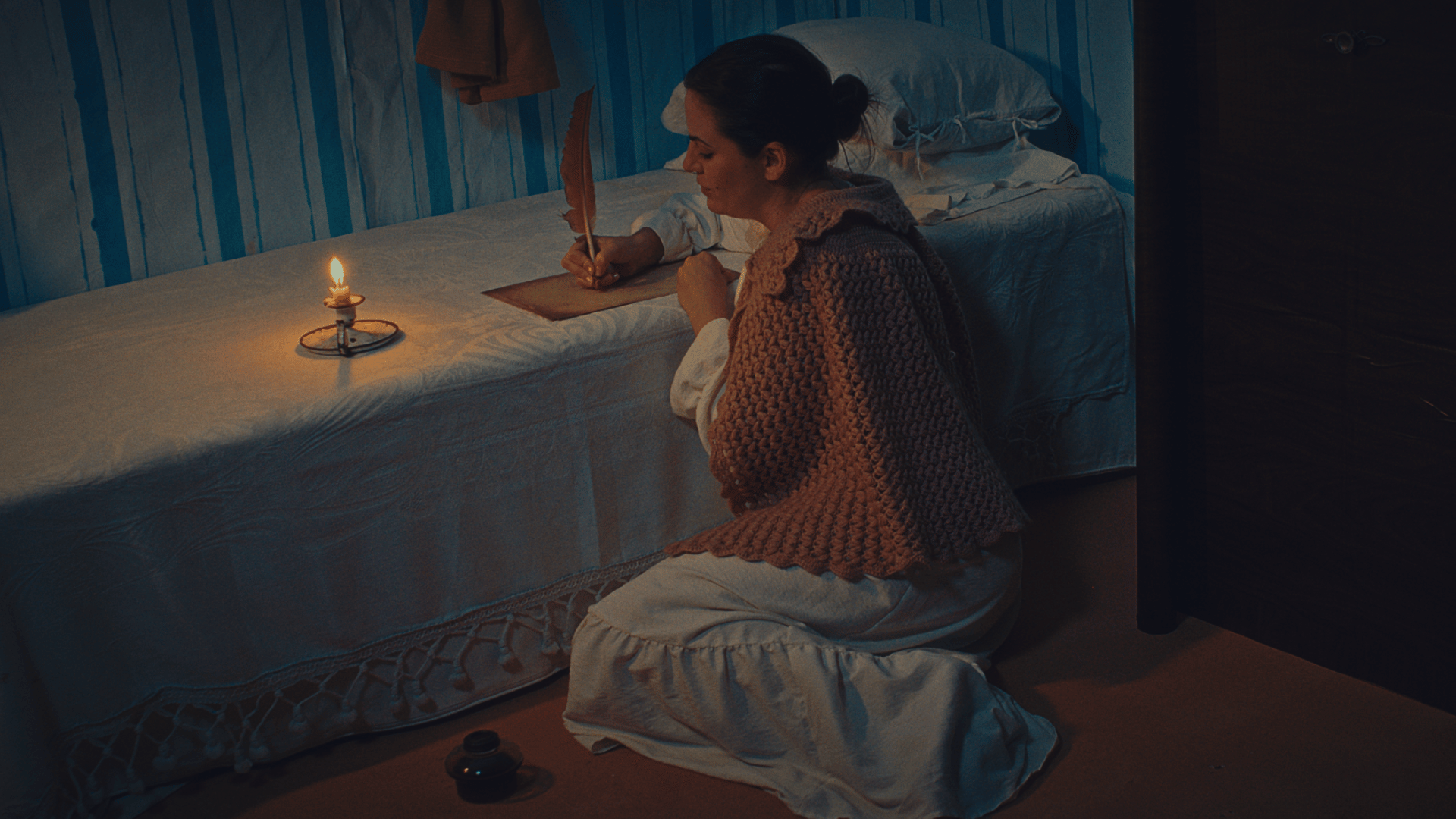 A imagem mostra uma mulher sentada no chão, debruçada sobre uma mesa escrevendo em um papel à luz de vela