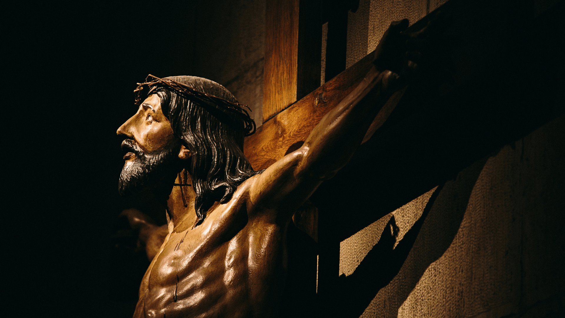 Escultura de Jesus crucificado na cruz, com coroa de espinhos, iluminada em ambiente escuro, destacando a expressão de sofrimento.
