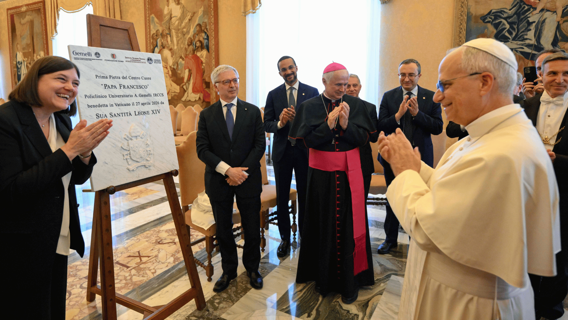 A imagem ilustra o Papa Leão XIV, com suas habituais vestes pontifícias, diante de uma placa em homenagem ao Papa Francisco. Ele está cercado posroutras pessoas e todos estão aplaudindo.