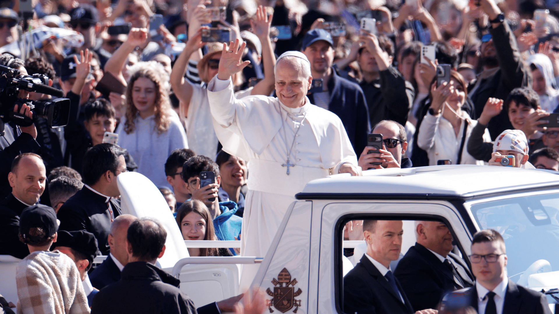 A imagem ilustra o Papa Leão XIV, com suas habituais vestes pontifícias, sobre o papamóvel acenando para os fiéis na Praça São Pedro