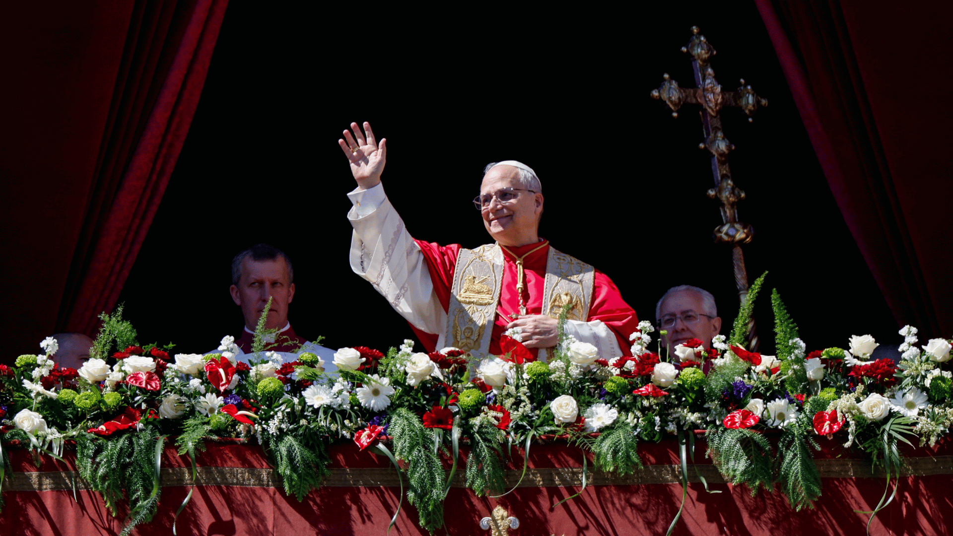 Papa Leão XIV acena para os fiéis do balcão central da Basílica de São Pedro A imagem ilustra o Papa Leão XIV, paramentado com a mozeta e uma estola branca com detalhes dourados, acenando para os fiéis com a mão direita erguida do balcão central da Basílica de São Pedro.
