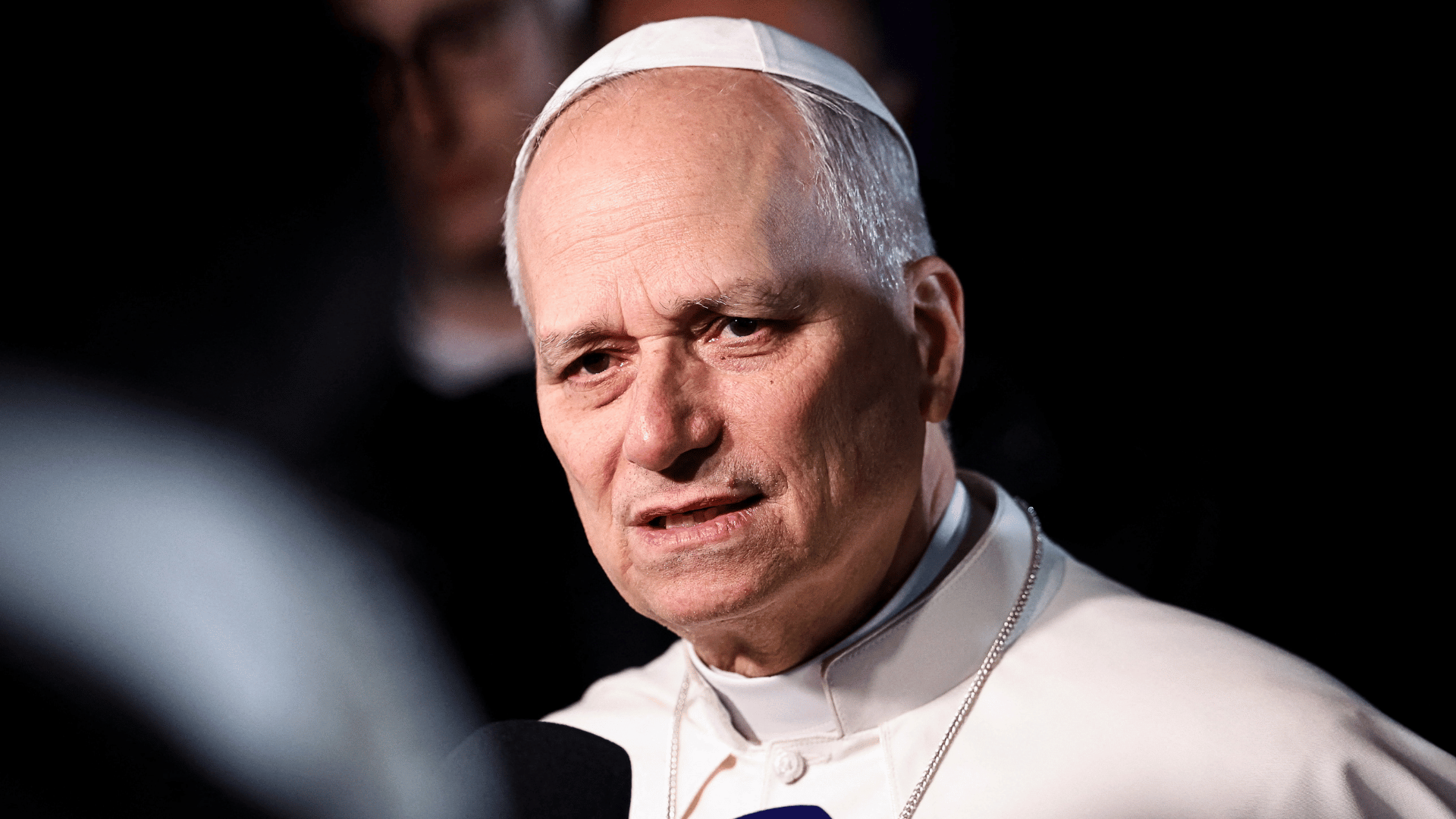 Papa conversa com jornalistas durante entrevista, com expressão séria e microfones em primeiro plano.