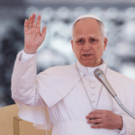 Papa levanta a mão enquanto fala ao microfone durante uma catequese.