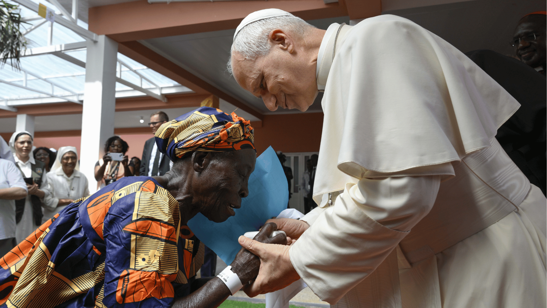 Papa Leão XIV cumprimenta e segura as mãos de uma pessoa idosa durante visita a centro de acolhimento.