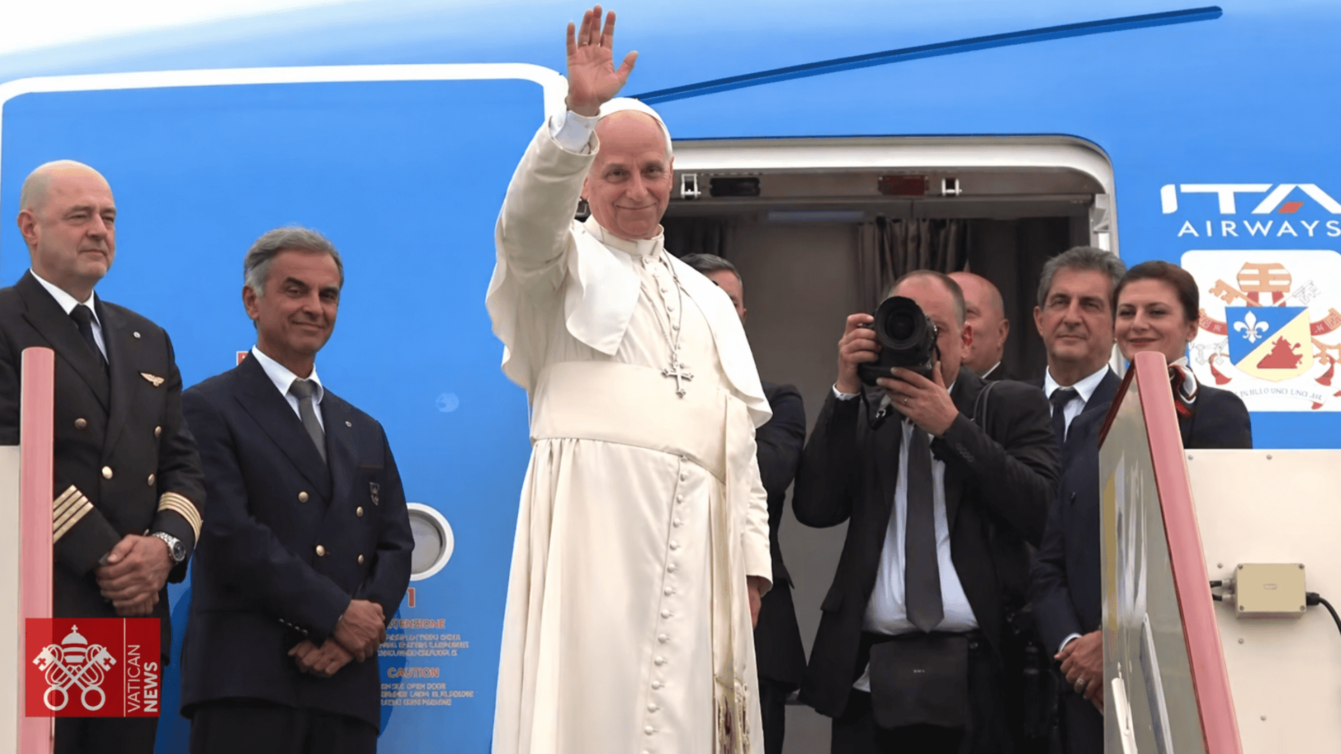 Papa Leão XIV acena na porta de avião antes da partida, cercado por integrantes da tripulação e equipe oficial.