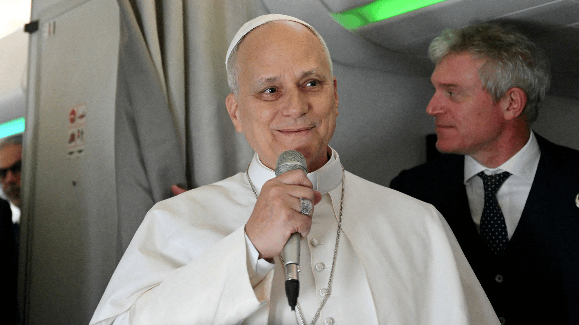 A imagem ilustra o Papa Leão XIV dentro de um avião, segurando um microfone com a mão direita.