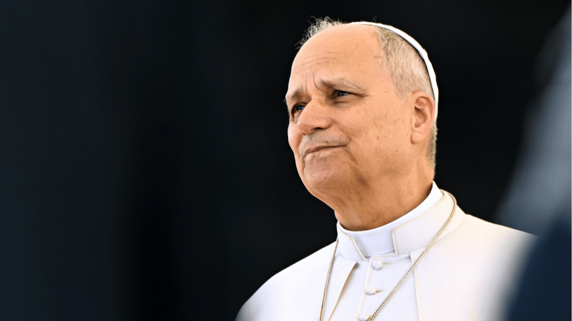 Papa Leão XIV com veste branca e solidéu, olhando para o lado diante de fundo escuro desfocado.