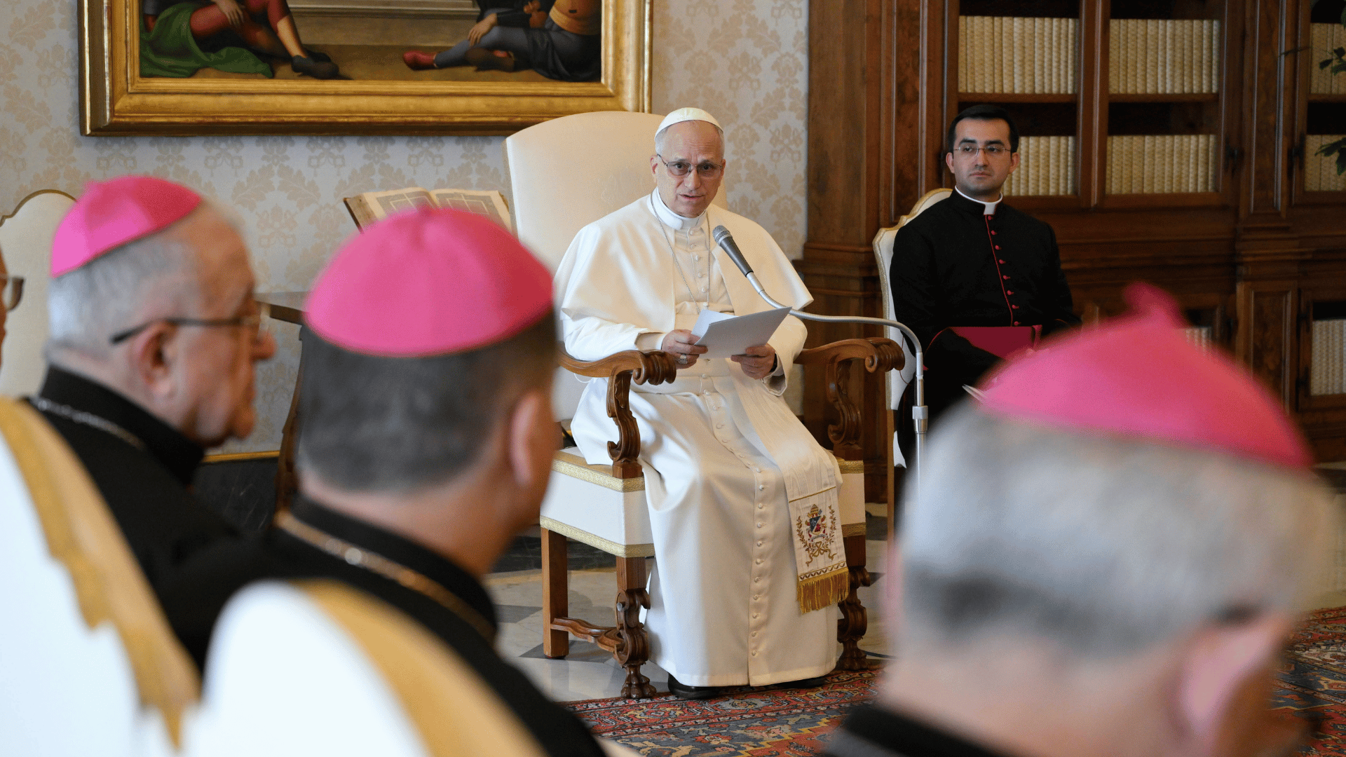 Papa sentado em cadeira fala ao microfone enquanto lê um texto, diante de bispos com solidéu rosa reunidos em sala com estantes de livros.