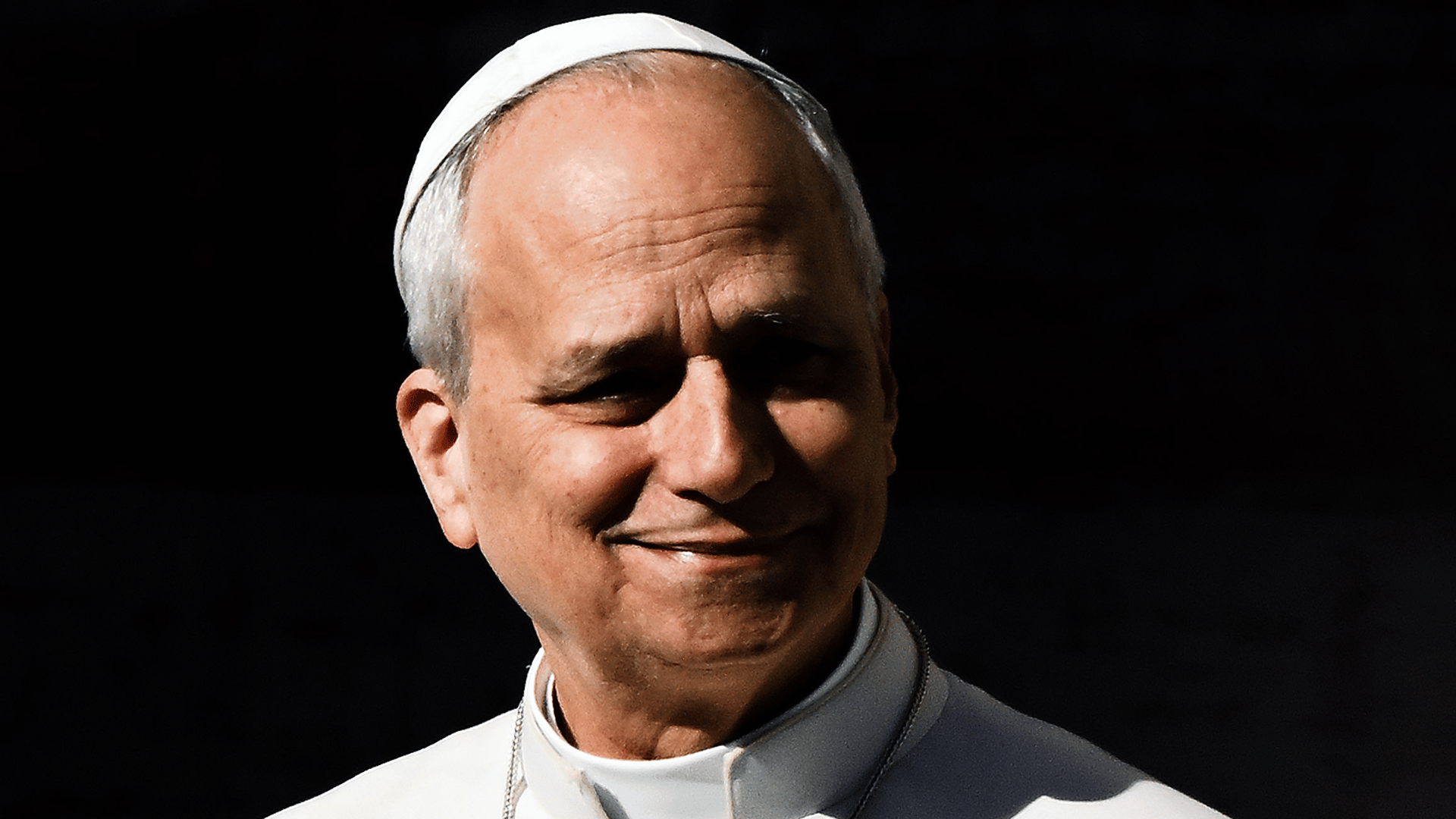Papa sorrindo, vestido de branco, com solidéu e expressão serena, sobre fundo escuro.