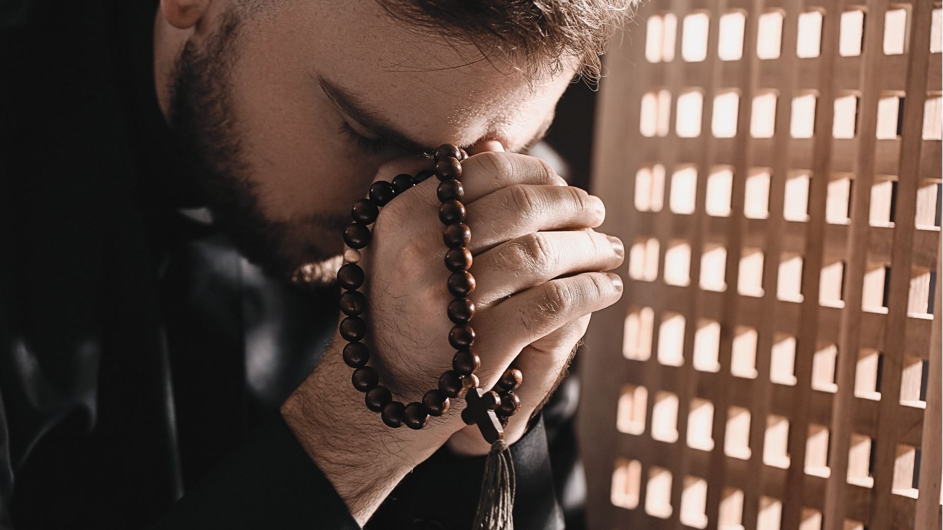 sacerdote dentro do confessionário com as mãos postas, contrito e segurando o terço
