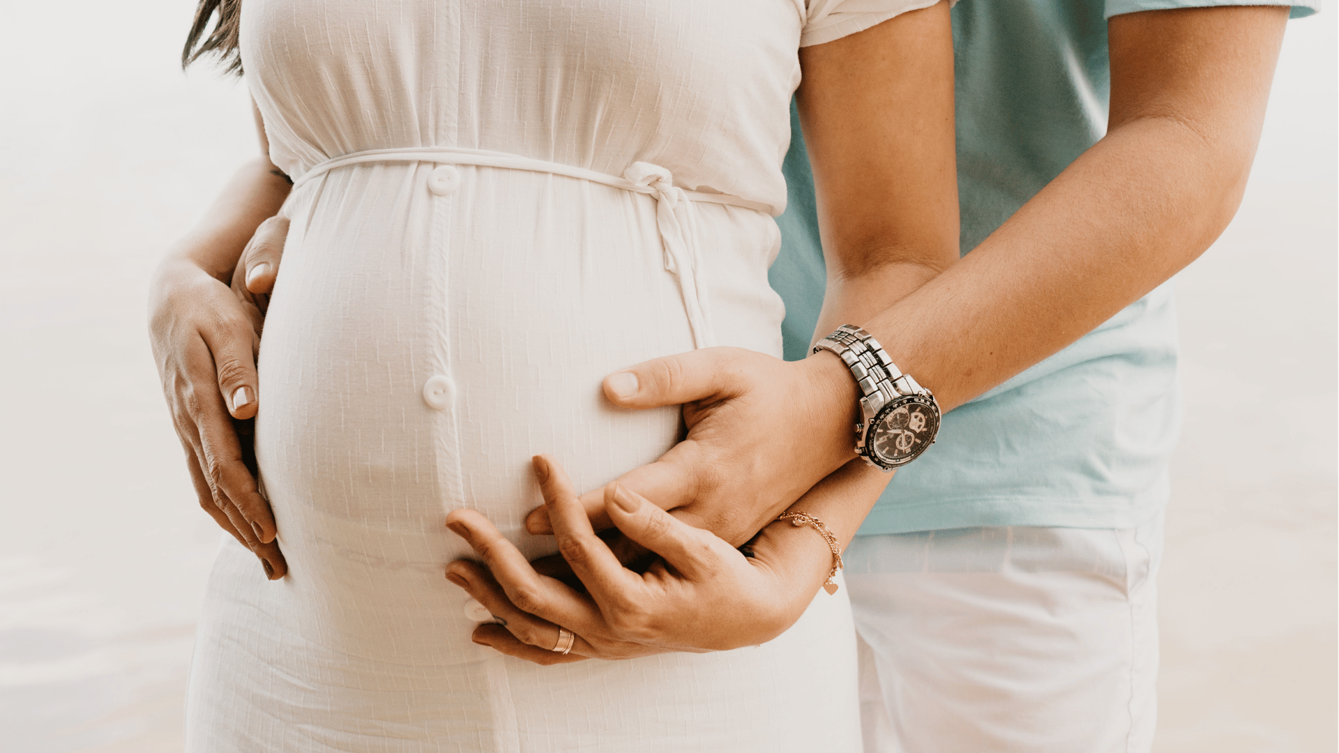 Casal em ensaio ao ar livre. A mulher é gestante, veste roupa clara enquanto o homem a abraça por trás, com as mãos sobre a barriga dela.