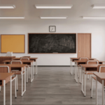 Sala de aula vazia com carteiras organizadas em fileiras, quadro negro ao fundo, mesa do professor, relógio na parede e janelas que iluminam o ambiente.