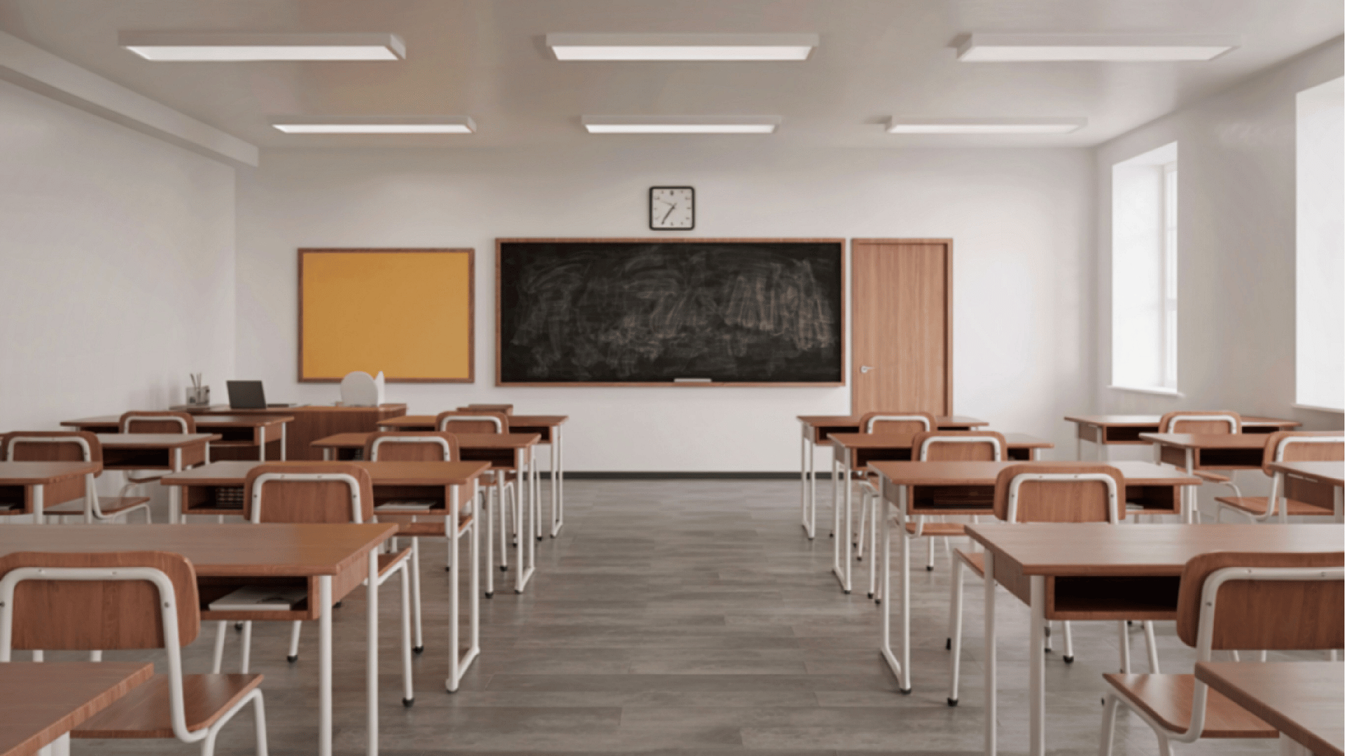 Sala de aula vazia com carteiras organizadas em fileiras, quadro negro ao fundo, mesa do professor, relógio na parede e janelas que iluminam o ambiente.
