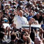 Papa acena para uma multidão de fiéis durante audiência geral desta quarta-feira, 20. Cercado por pessoas que registram o momento com celulares, o Pontífice aparece em pé no papamóvel saudando os presentes.