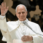 Papa acena aos participantes durante encontro com a Renovação Carismática Católica na Sala Paulo VI, no Vaticano. Diante de um microfone, ele sorri enquanto saúda os fiéis.