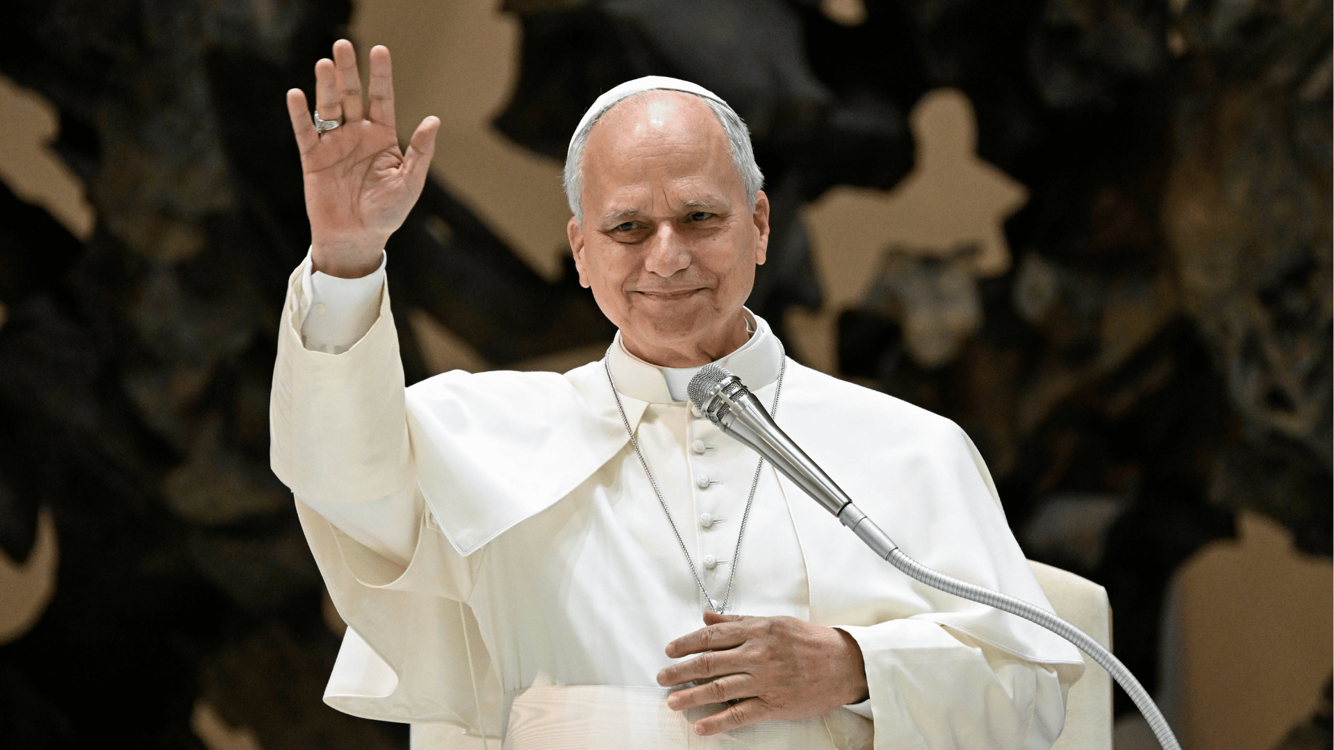 Papa acena aos participantes durante encontro com a Renovação Carismática Católica na Sala Paulo VI, no Vaticano. Diante de um microfone, ele sorri enquanto saúda os fiéis.