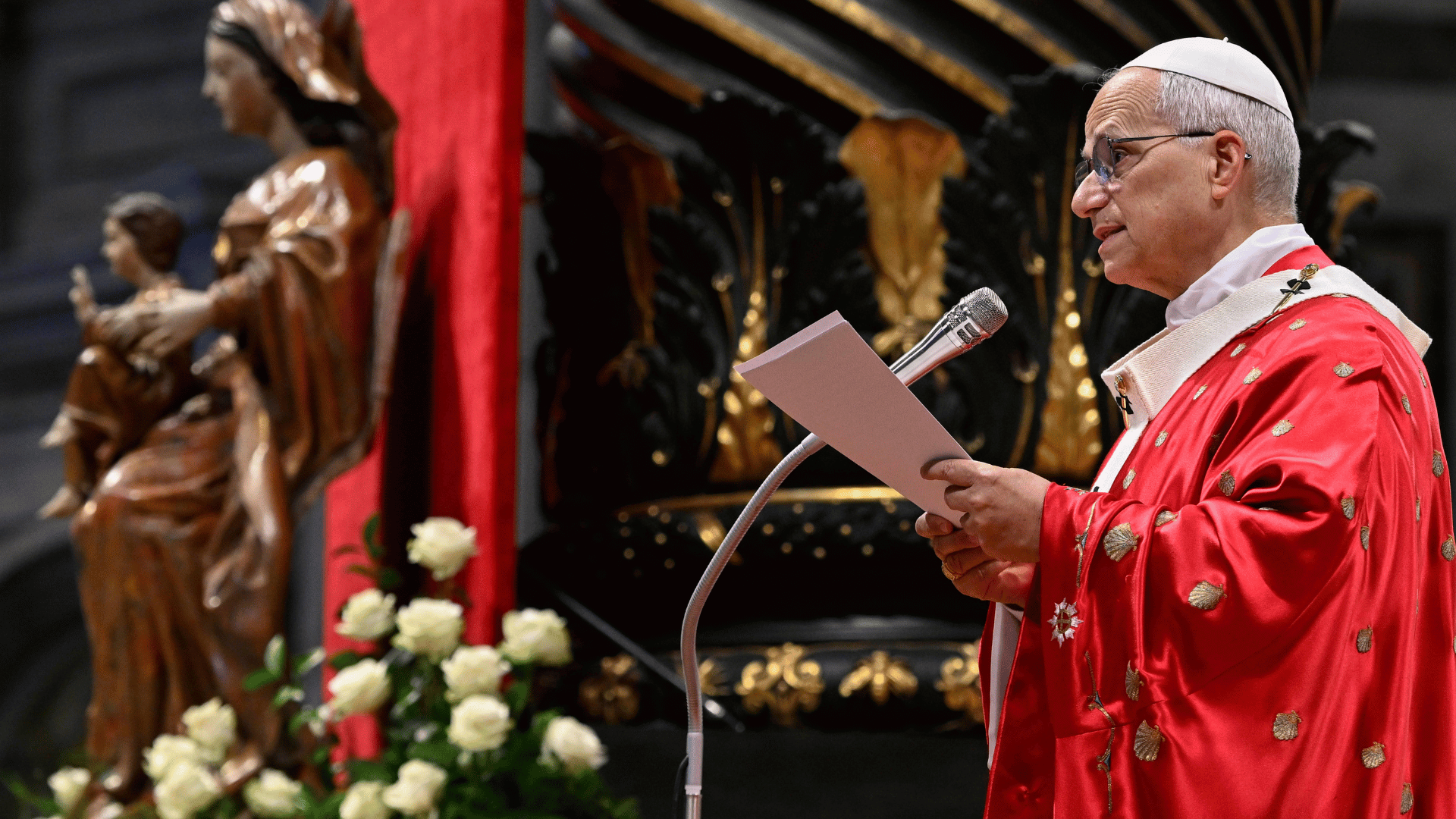 O Papa Leão XIV aparece paramentado com vestes litúrgicas vermelhas e fala ao microfone segurando um papel em suas mãos