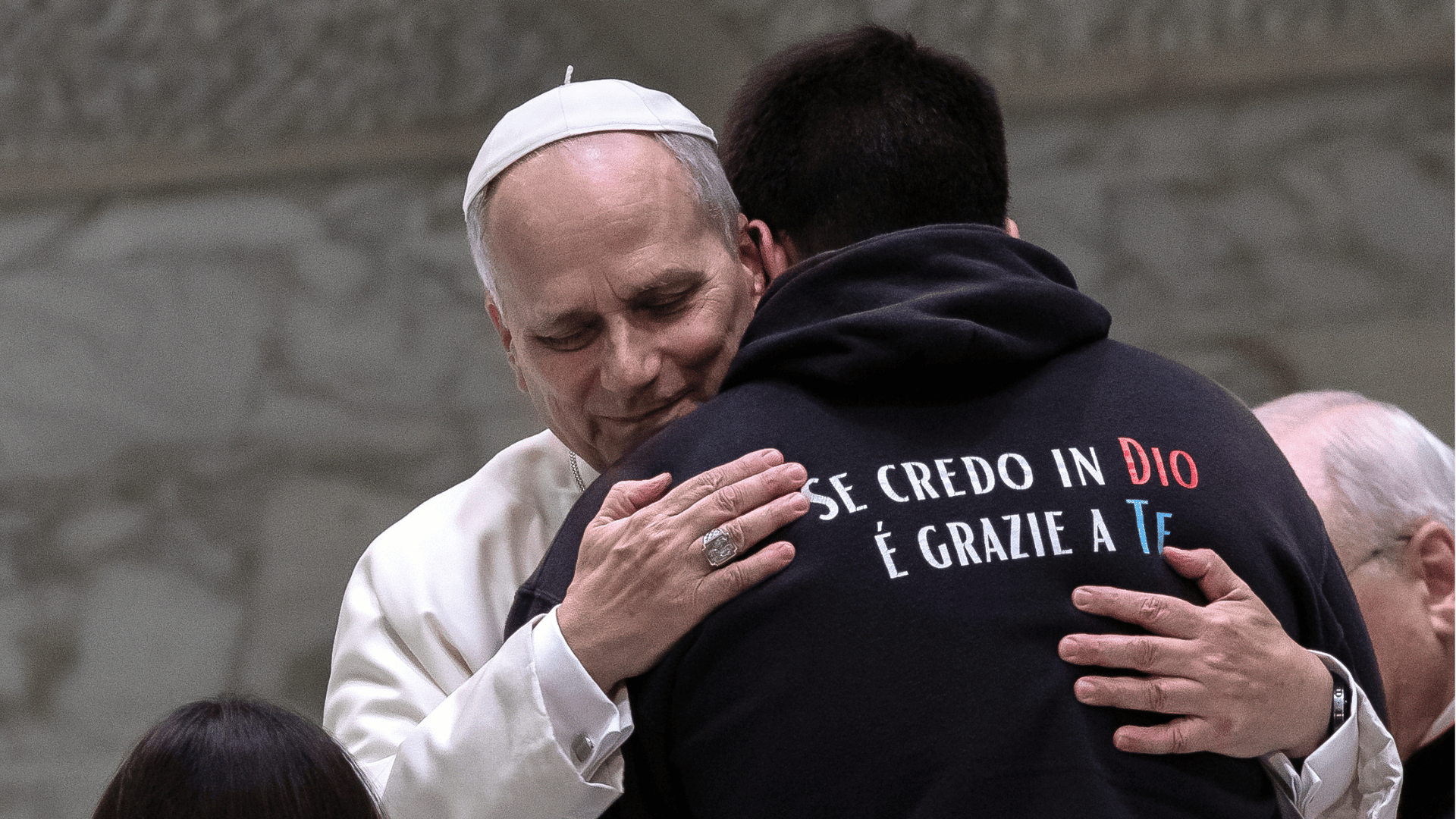 Papa abraça um jovem de costas, que usa moletom com a frase “Se creio em Deus é graças a você”, na Sala Paulo VI, no Vaticano.