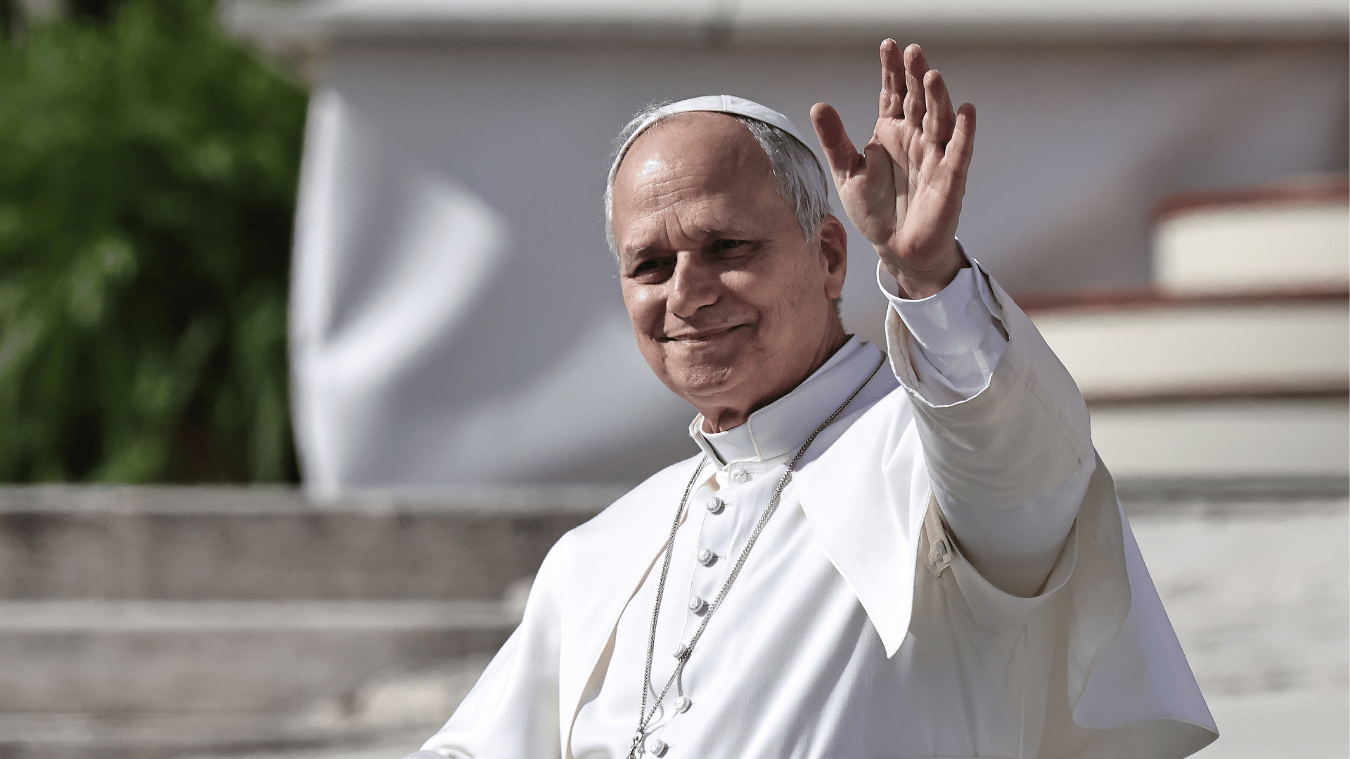 A imagem ilustra o Papa Leão XIV, com suas habituais vestes pontifícias, com a mão esquerda erguida em gesto de aceno.
