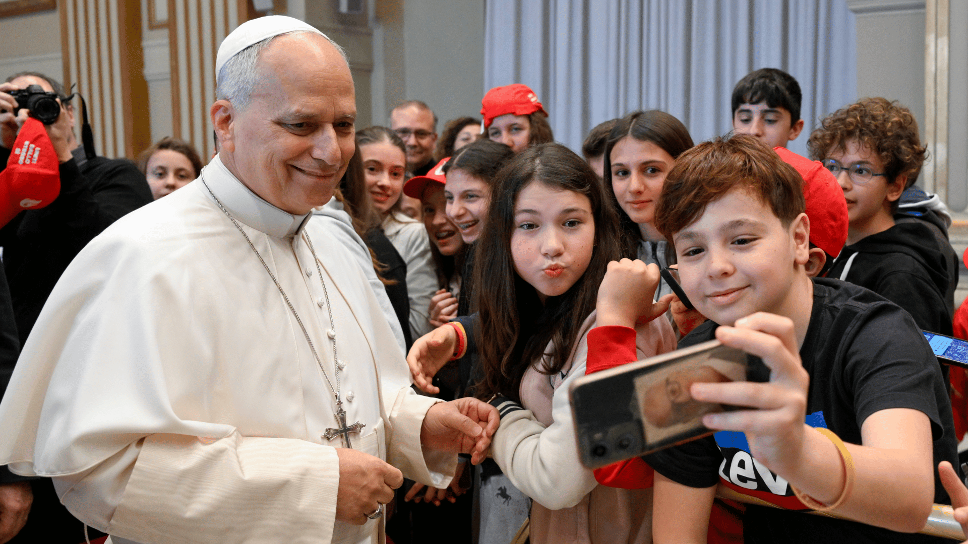 A imagem ilustra o Papa Leão XIV, com suas habituais vestes pontifícias, tirando uma selfie com dois adolescentes, um menino e uma menina, cercados por outros jovens.