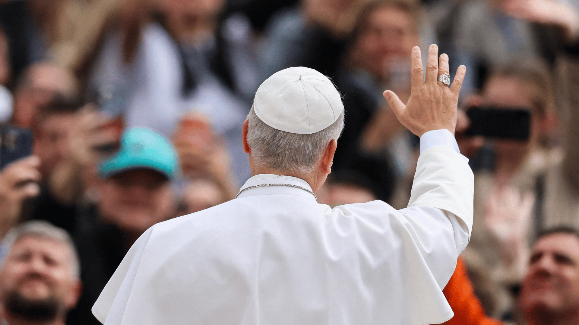 A imagem ilustra o Papa Leão XIV de costas, com o braço direito erguido em um aceno para os fiéis que estão no fundo da foto.