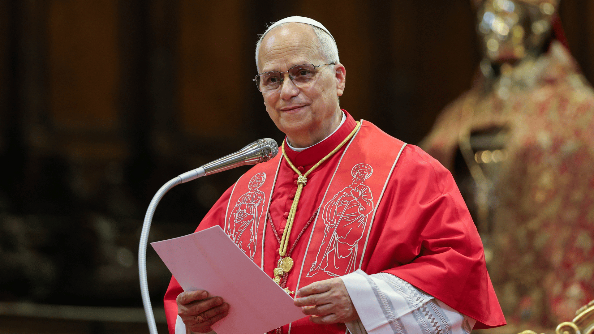 A imagem ilustra o Papa Leão XIV, paramentado com a mozeta e uma estola vermelha, discursando. Ele está diante de um microfone e tem algumas folhas de papel em suas mãos.