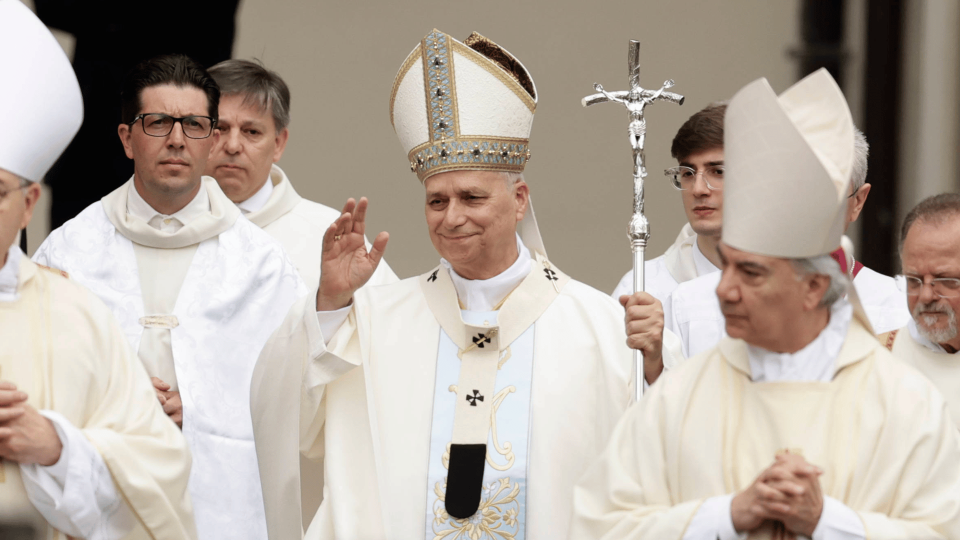 A imagem ilsutra o Papa Leão XIV, paramentado com vestes litúrgicas brancas, carregando a férula papal com a mão esquerda e acenando com a direita. Ao redor dele estão alguns bispos e padres.