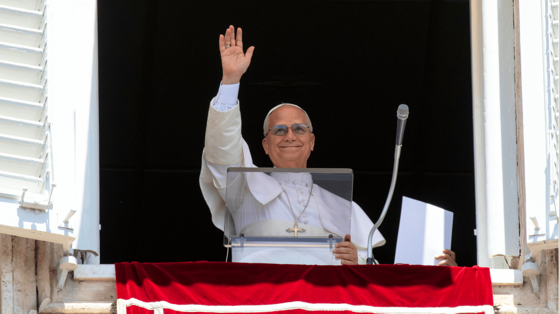 Papa aparece na janela do Vaticano acenando aos fiéis durante oração do Regina Caeli. Vestido de branco, ele sorri enquanto faz a saudação, ao lado de um microfone e de uma bandeira vermelha com o brasão da Santa Sé na sacada.