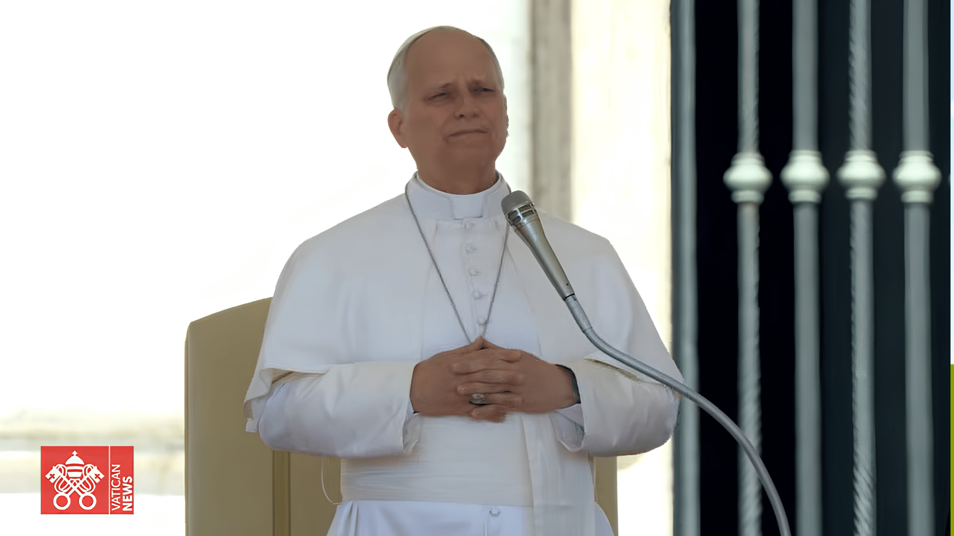 A imagem mostra o Papa Leão XIV em pé, com suas tradicionais vestes brancas, aos mãos cruzadas à sua frente, na altura da cintura, em posição de oração. O Papa está atrás de um microfone. 