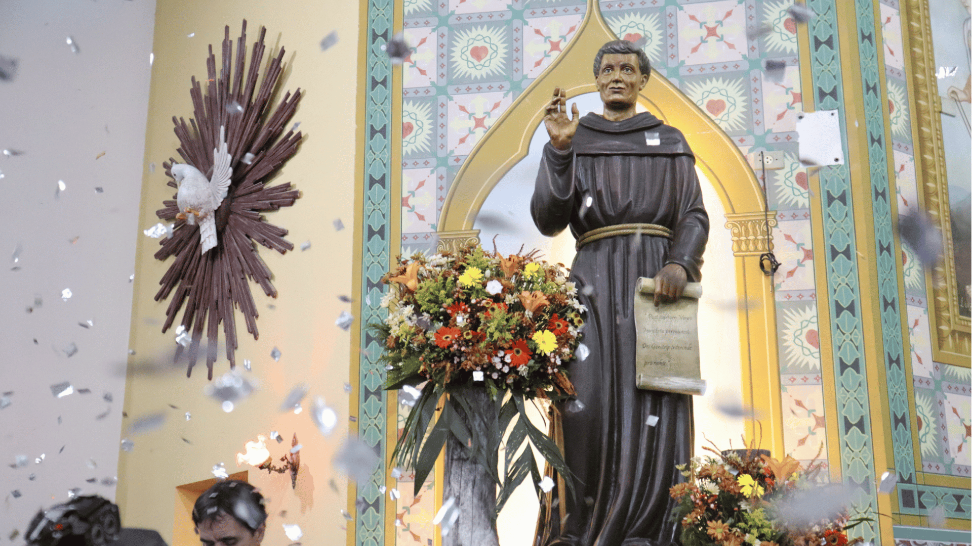 Imagem de Frei Galvão no Santuário dedicado a ele, em Guaratinguetá (SP),  ornamentada com flores, enquanto confetes caem durante celebração festiva. Ao lado da imagem, aparece o símbolo do Espírito Santo em forma de pomba.
