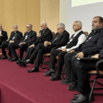 Sentados em um palco, os bispos participantes acompanham as discussões enquanto um dos bispos faz uso da palavra. Ao fundo, há elementos litúrgicos e uma imagem religiosa, compondo o ambiente do encontro pastoral.