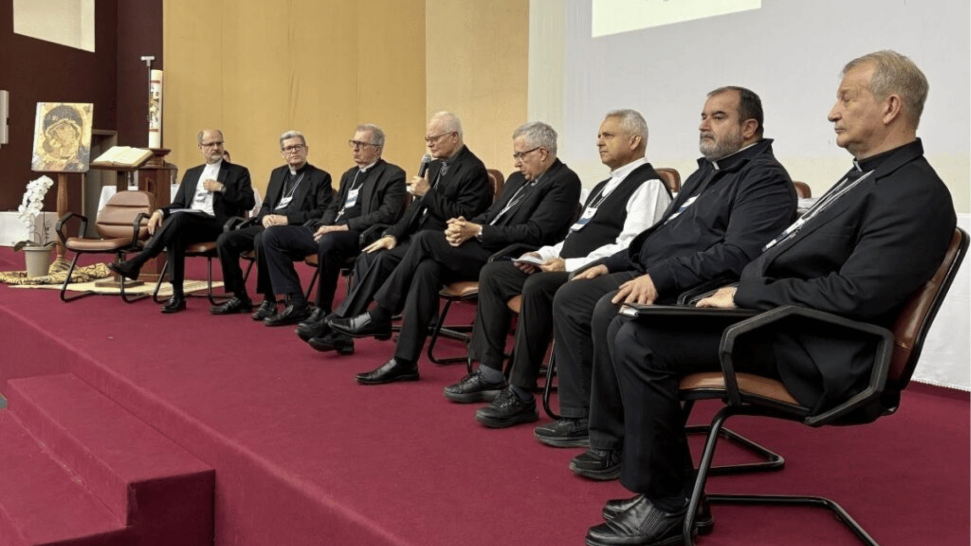 Sentados em um palco, os bispos participantes acompanham as discussões enquanto um dos bispos faz uso da palavra. Ao fundo, há elementos litúrgicos e uma imagem religiosa, compondo o ambiente do encontro pastoral.