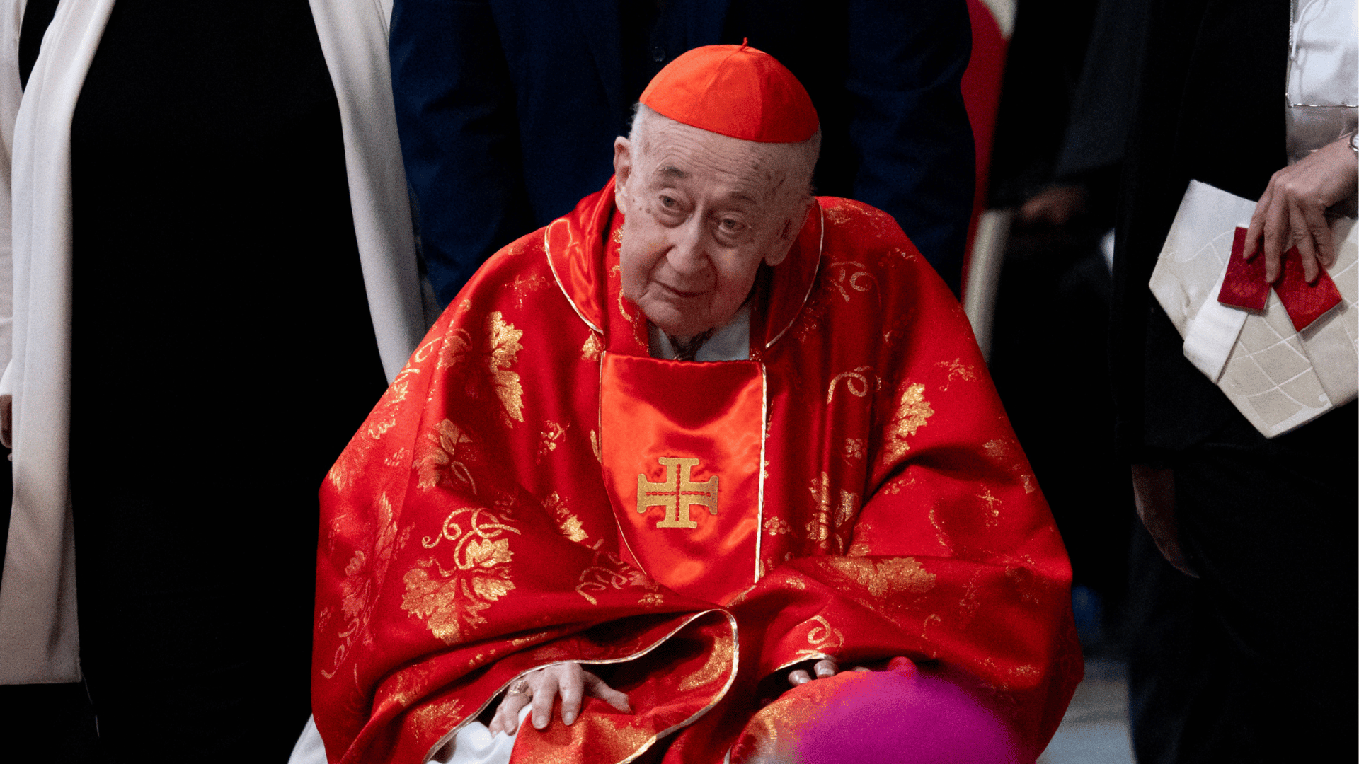 Cardeal Camillo Ruini, vestido com paramentos vermelhos litúrgicos e solidéu cardinalício, participa de uma celebração religiosa. Sentado e cercado por outras pessoas, ele aparece em destaque no centro da imagem, com expressão serena.